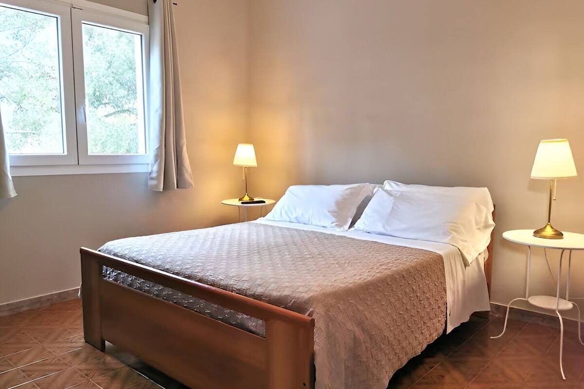 A neatly made bed with white linens, two bedside lamps, and a window with curtains.