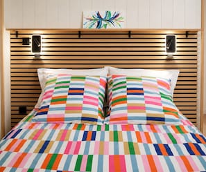 A bed with a colorful striped bedspread and pillows, a wooden slatted headboard, and two wall-mounted speakers.