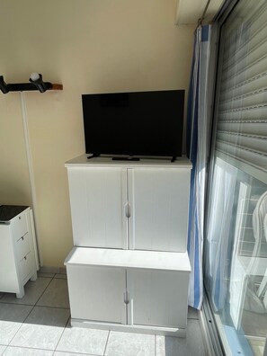 Une pièce avec un téléviseur posé sur un meuble, une porte-fenêtre coulissante et un meuble blanc avec des tiroirs.