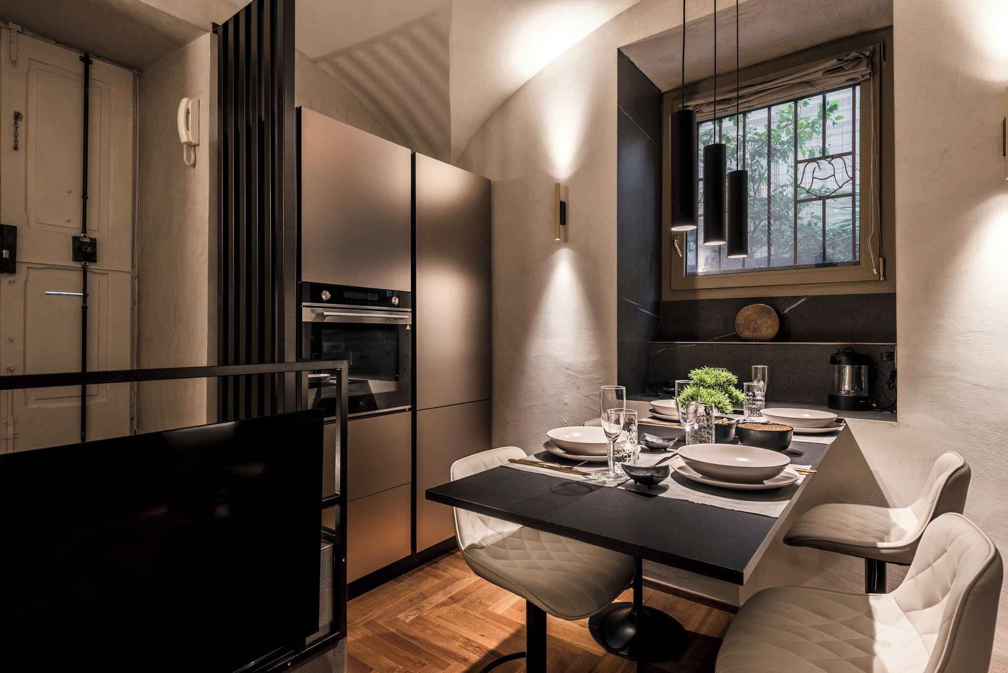 A modern kitchen with a dining area, featuring a large flat-screen TV, a dining table set for four, and a window with a view of greenery.