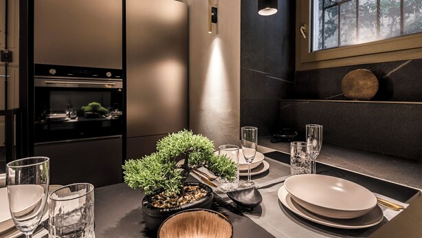 Cozinha moderna com eletrodomésticos embutidos, área de jantar com mesa posta para dois e vista para árvores pela janela.