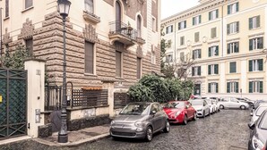 Uma rua de calçada estreita, flanqueada por carros estacionados e edifícios históricos com sacadas e persianas.