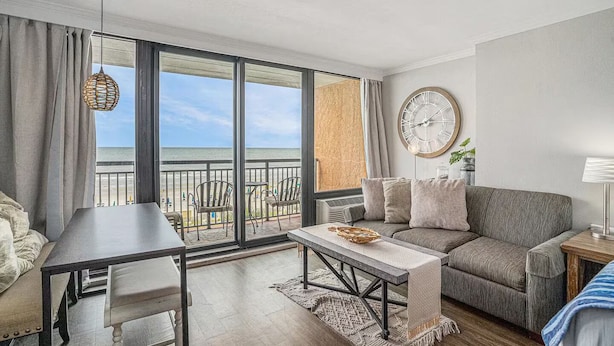 A modern living room with a balcony overlooking the beach, a round wall clock, and a dining area with a table and chairs. - Cozy condo in wonderful Myrtle Beach with refreshing AC (Myrtle Beach)