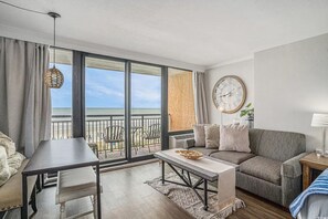 Uma sala de estar moderna com uma varanda com vista para a praia, um relógio de parede redondo e uma área de jantar com mesa e cadeiras.