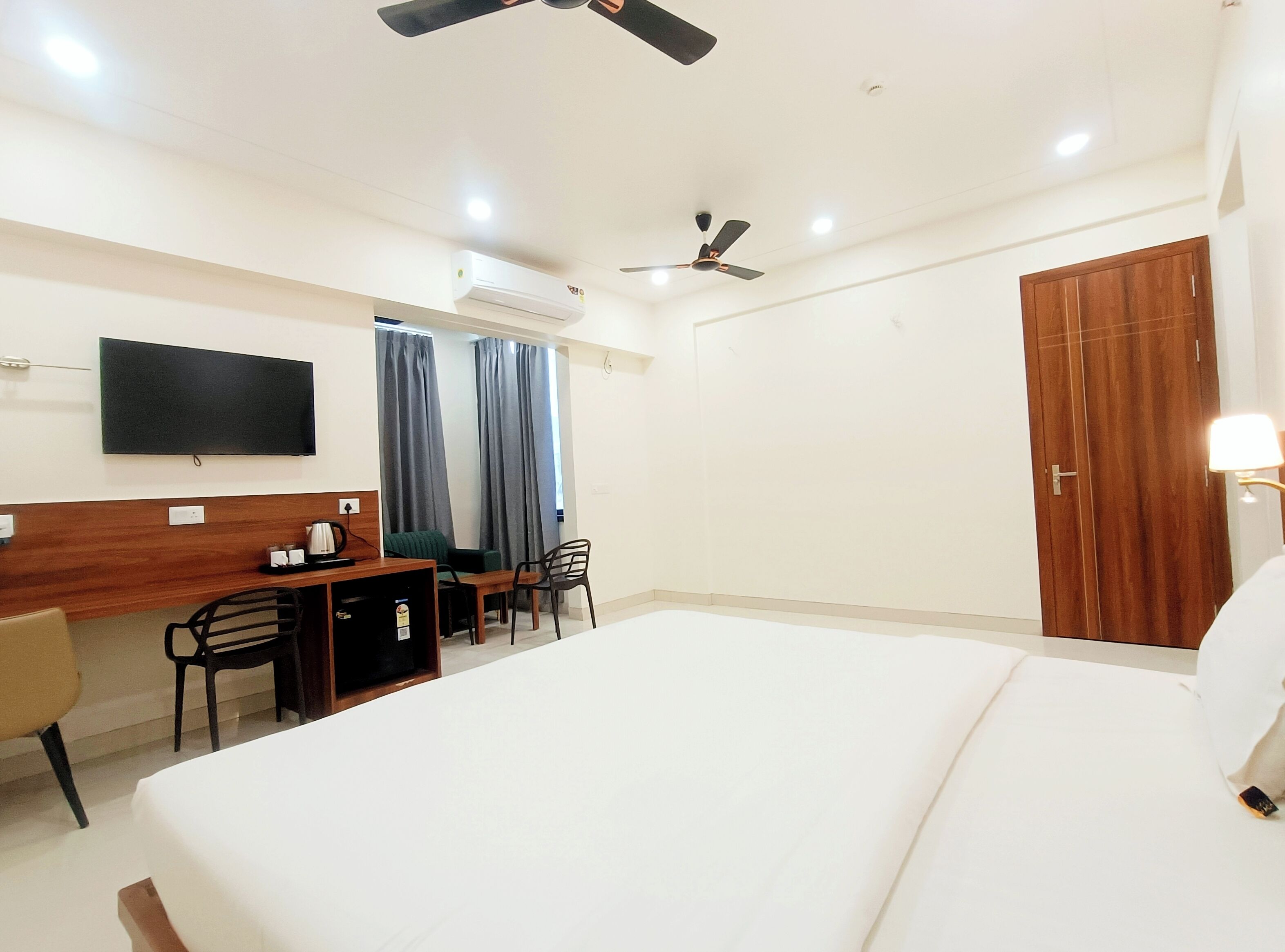A hotel room with a bed, a desk with chairs, a television, a wardrobe, and two ceiling fans.