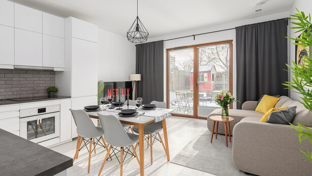 A modern kitchen with white cabinets, a dining area with a table and chairs, a sofa with cushions, and a sliding glass door leading to a balcony. - Lumina Family apartment with patio (Warsaw)