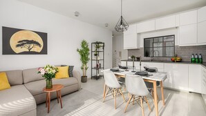 A modern living room with a sofa, a dining table with chairs, a potted plant, and a painting of a tree against a moon. - Lumina Family apartment with patio (Warsaw)
