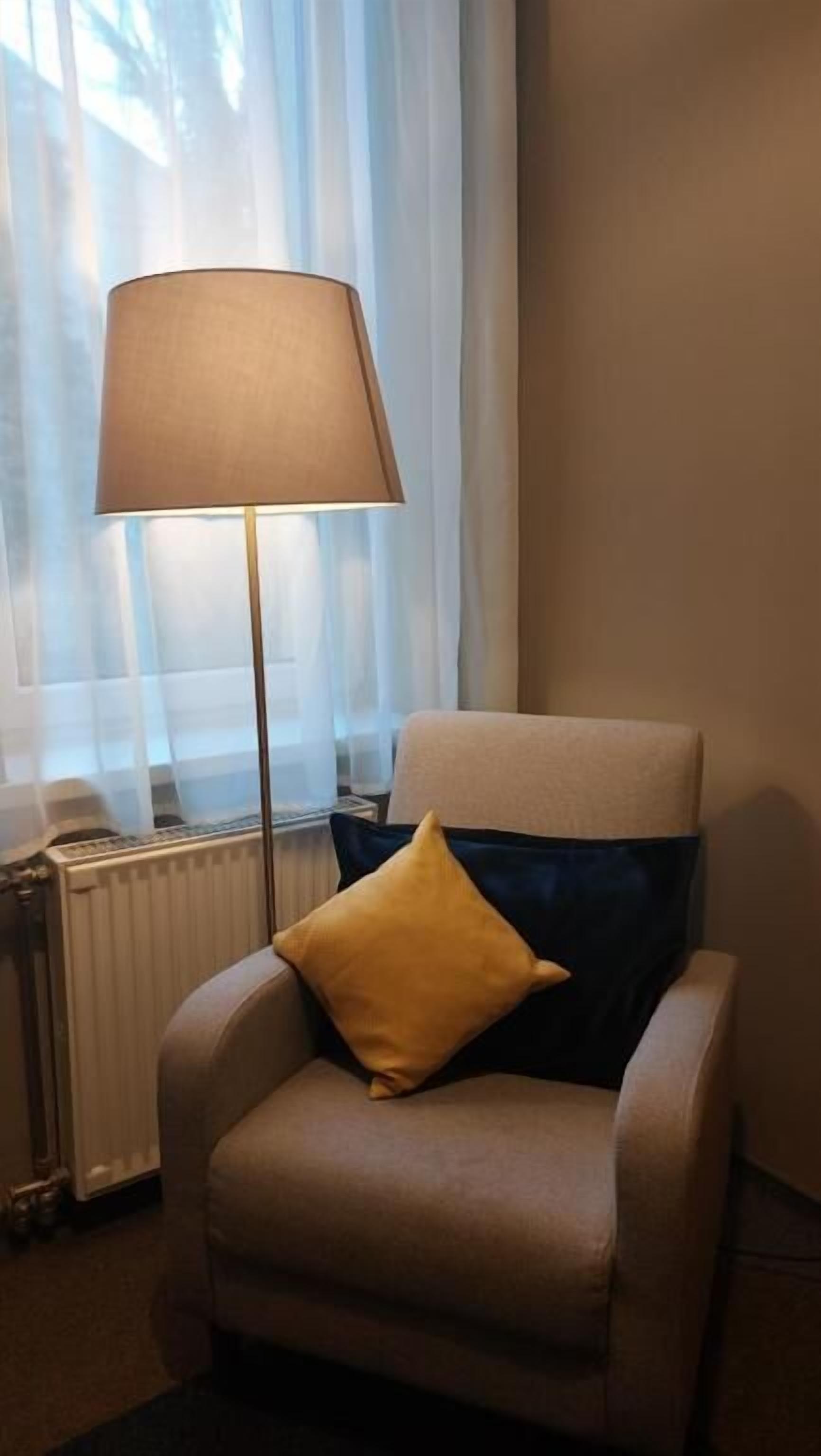 Une chaise avec un coussin, un lampadaire et un radiateur dans une pièce où sont accrochées des rideaux légers.