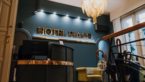 Un mostrador de recepción de hotel con un cartel que dice "HOTEL PIANO", una araña, una barra de recepción y una zona de estar con una silla y una mesa.