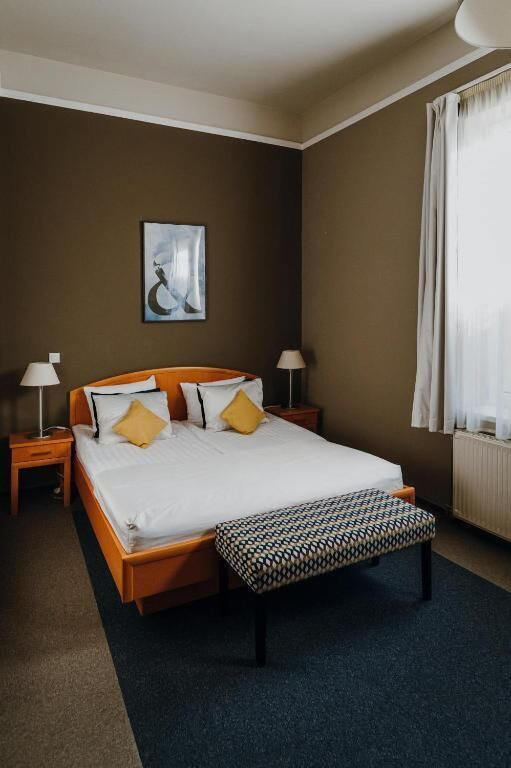 A neatly arranged hotel room with a large bed, two bedside tables with lamps, a bench, a framed artwork on the wall, and a window with curtains.