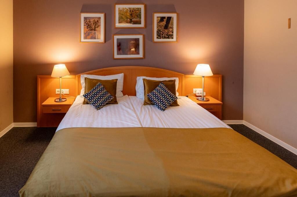 Een hotelkamer met een groot bed, twee nachtkastjes met lampen, en ingelijste foto’s aan de muur.
