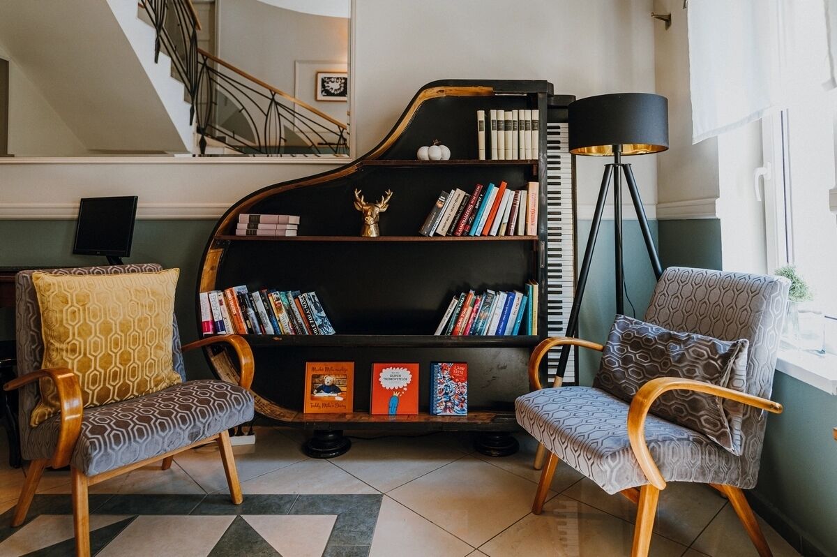A living room with a grand piano, two patterned chairs, a bookshelf, and a floor lamp.