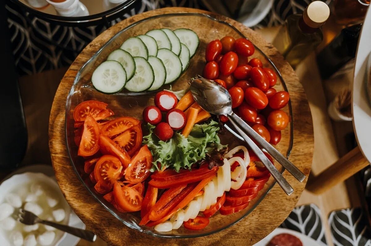 A glass bowl filled with sliced vegetables and cherry tomatoes on a wooden board.
