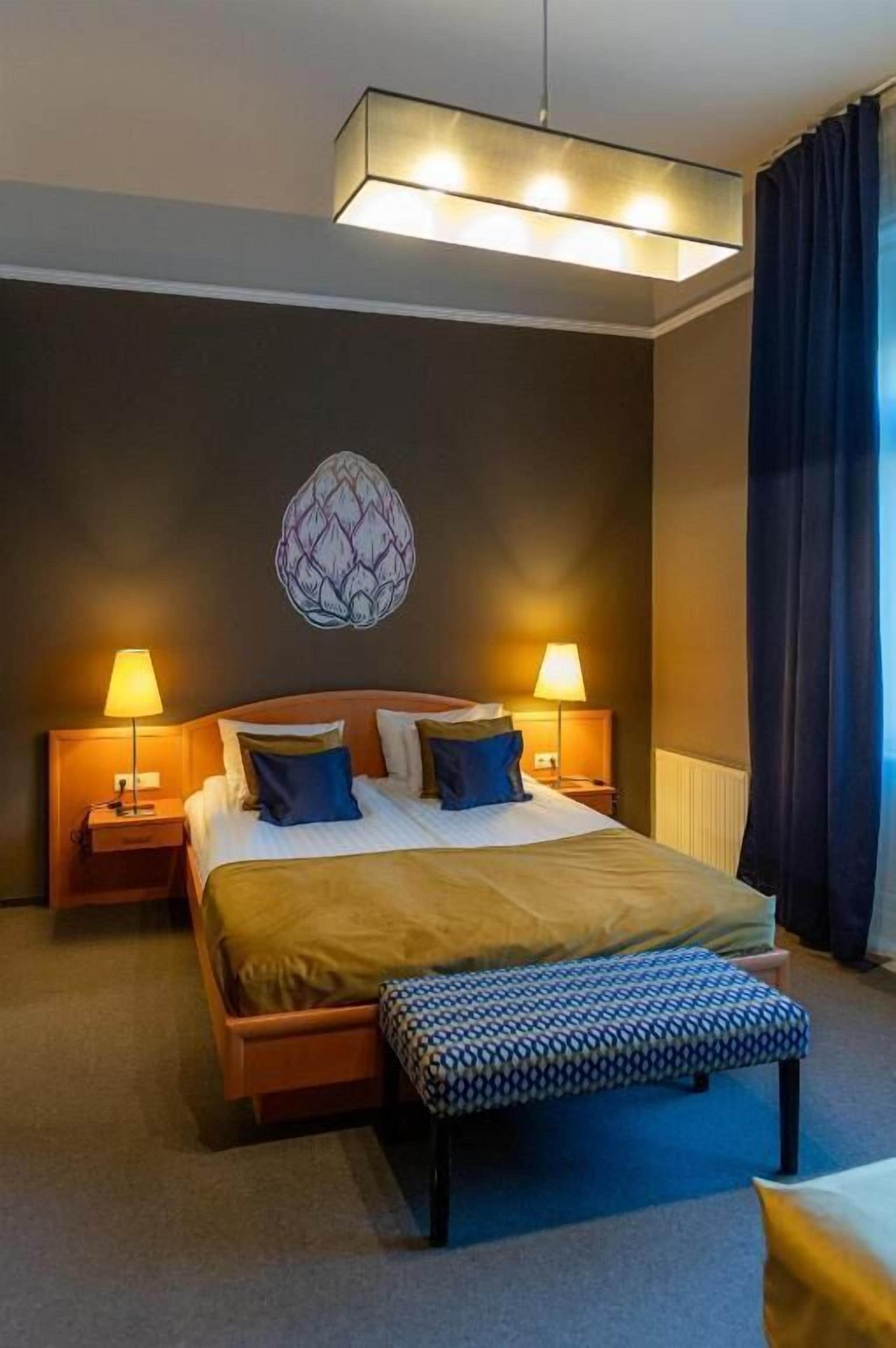 A hotel room with a large bed, two bedside lamps, a wall-mounted artwork, a bench, and a window with curtains.