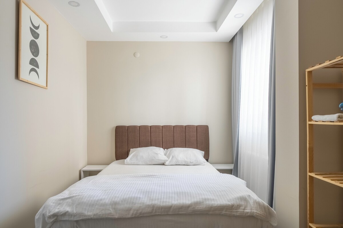 Un lit bien fait, avec une tête de lit, une table de chevet, une œuvre d’art accrochée au mur et une fenêtre avec des rideaux.