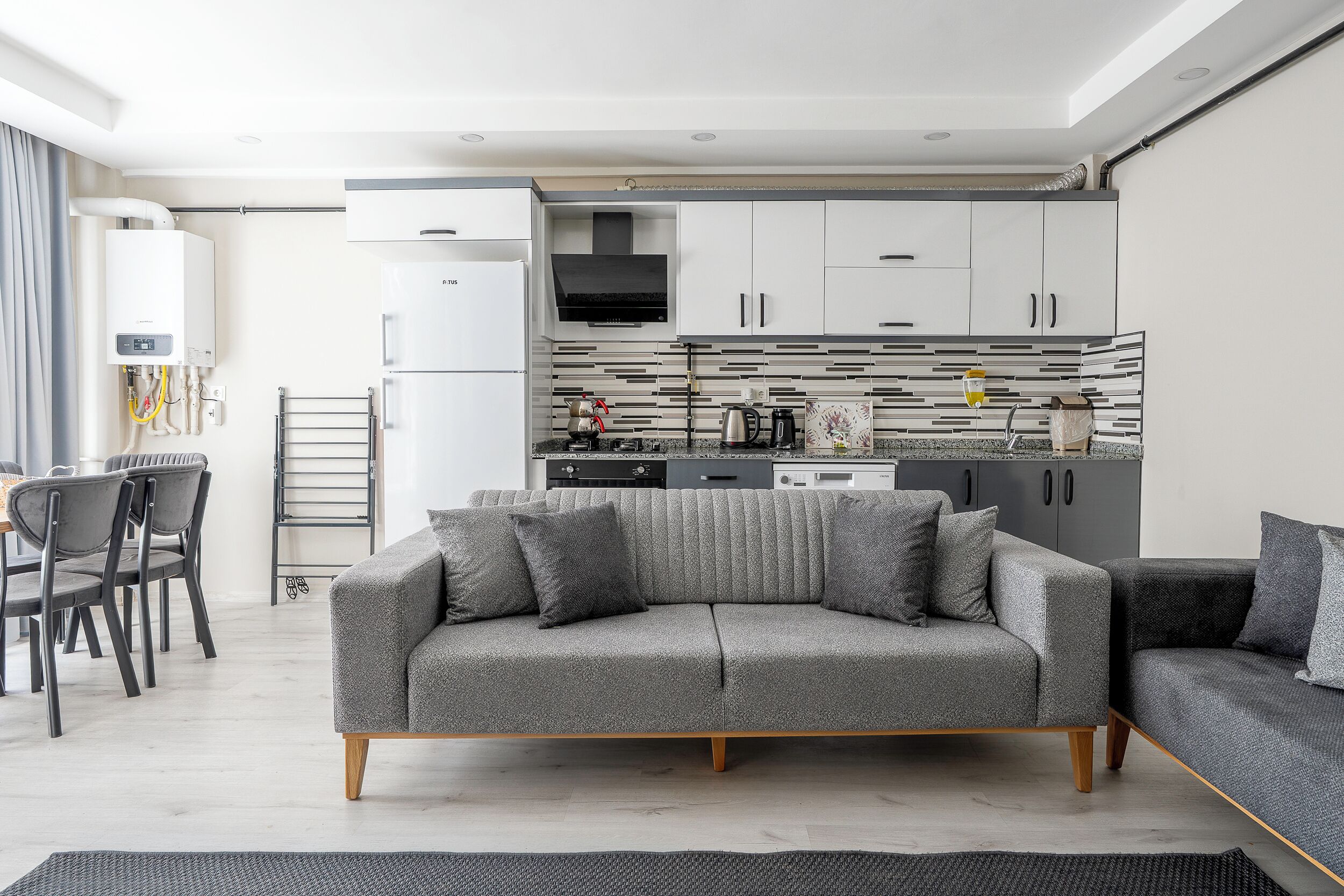 A modern living room with a grey sofa, a dining area with chairs, and a kitchen with white cabinets and a black range hood.