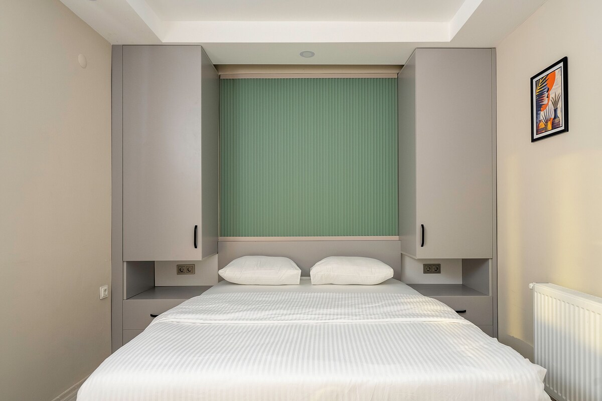 Une chambre à coucher avec un lit, deux tables de chevet, une fenêtre avec des stores verts et un tableau encadré au mur.