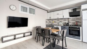 Une cuisine moderne dotée d’un four encastré, d’un coin repas avec une table et des chaises en bois, d’un téléviseur à écran plat fixé au mur et d’une fenêtre avec des rideaux.