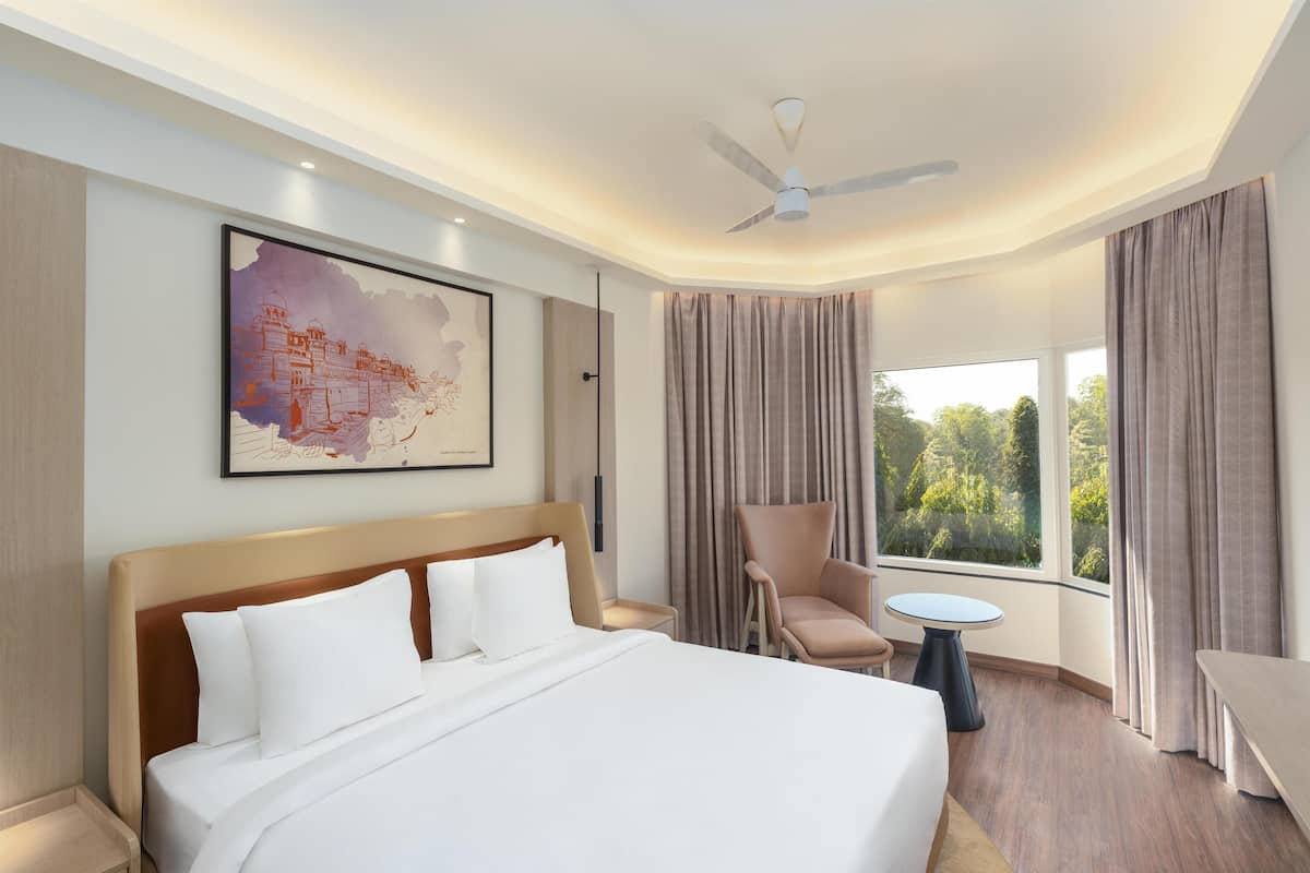 A hotel room with a large bed, a chair, a small table, a ceiling fan, and a window with curtains.