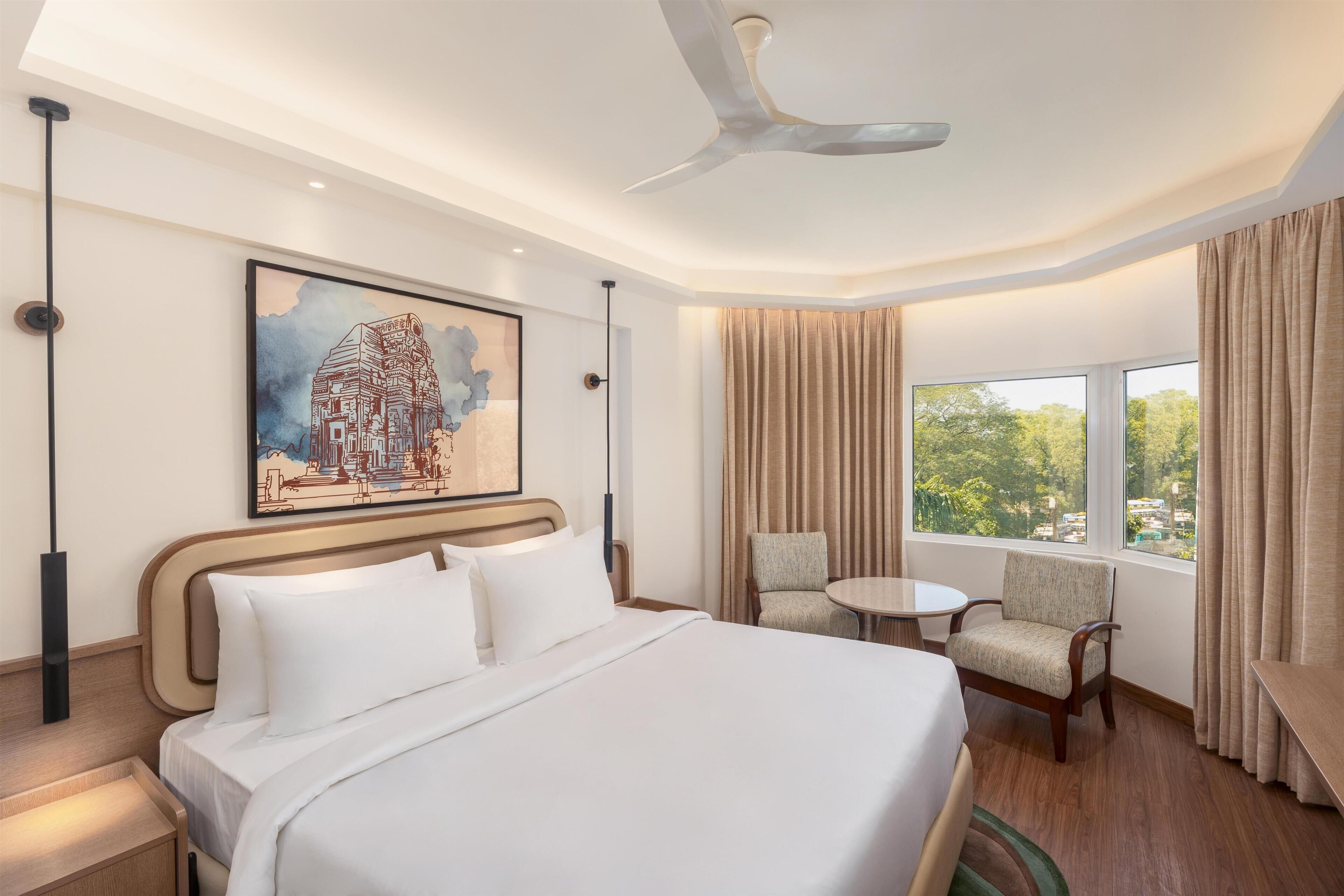 A hotel room with a large bed, a chair, a small table, a ceiling fan, and a window with curtains.