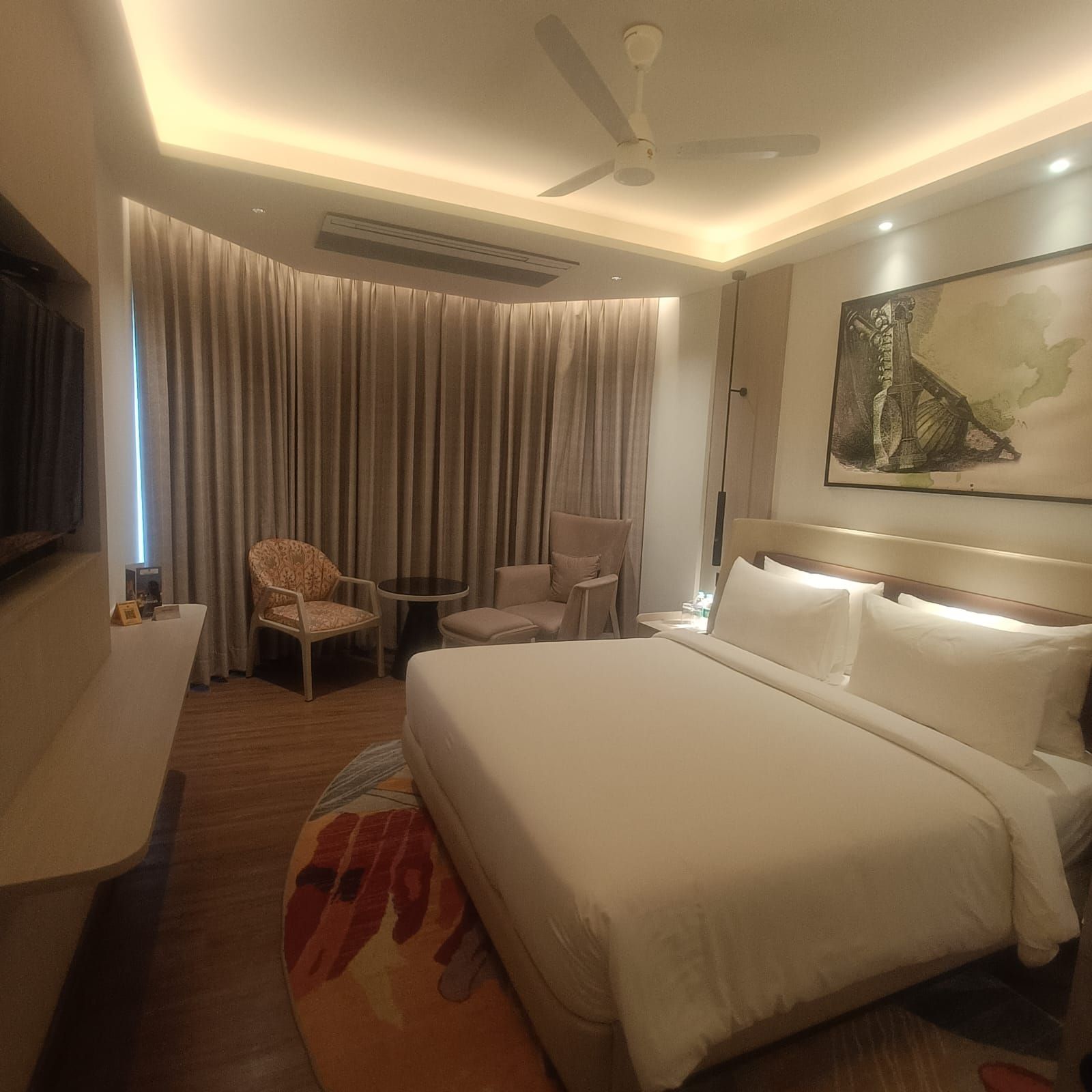 A hotel room with a large bed, a ceiling fan, a flat-screen TV, a small table, and a chair.