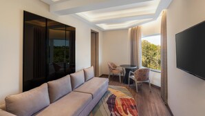 A modern hotel room with a sofa, a small table with chairs, a flat-screen TV, and a large window with curtains. - Hotel Gwalior Regency, a member of Radisson Individuals (Gwalior)