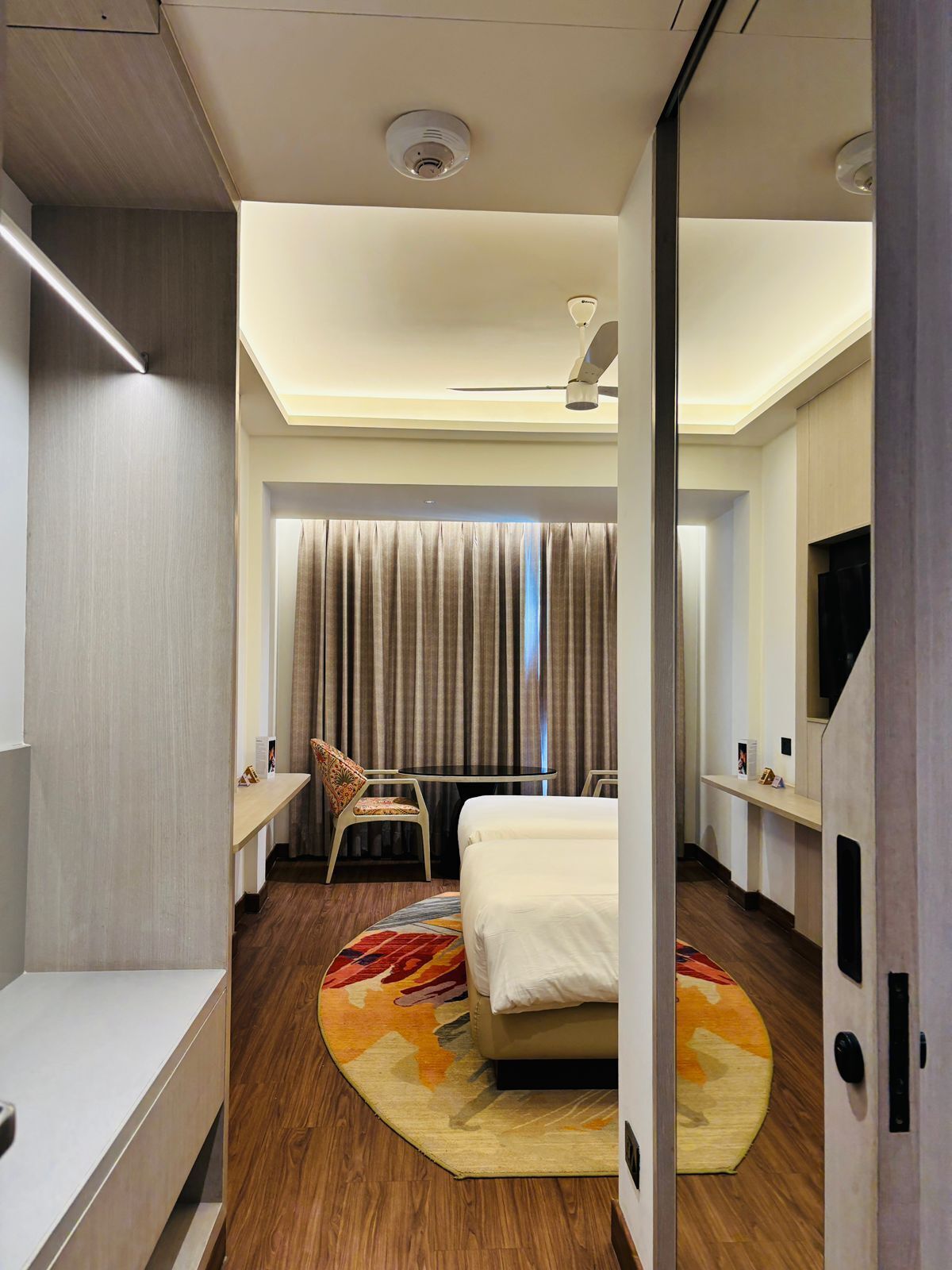 Habitación de hotel con cama, escritorio, silla, ventilador de techo y ventana con cortinas.