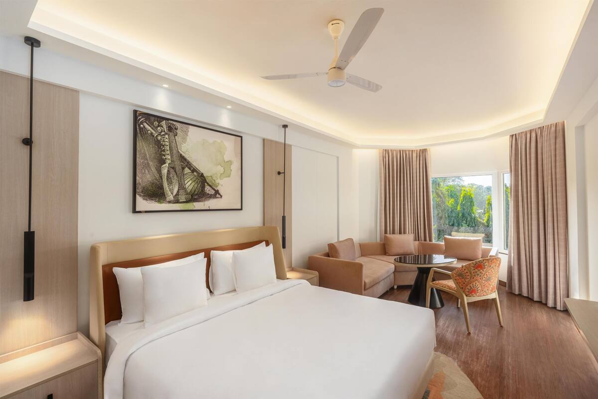 A modern hotel room with a large bed, a sofa, a chair, a small table, and a ceiling fan.