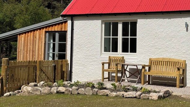 Una casa con techo rojo, paredes exteriores blancas, una chimenea y un pequeño patio con dos bancos de madera.