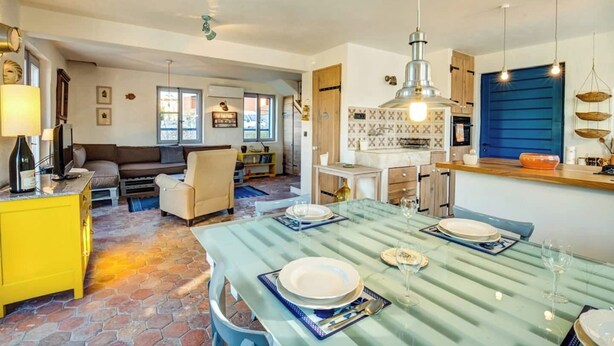 A dining area with a table set for four, a kitchen with blue cabinets, and a living room with a sofa and a TV. - Secluded stone villa with private pool on peaceful Solta Island (Gornje Selo)