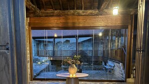 An interior space with a stone floor, a wooden table with a vase, and a view through large windows to an outdoor area with stone walls and a wooden deck. - Luxury suite in charming B&B in Galicia (O Agro)