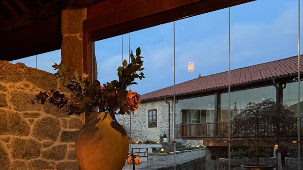 A stone wall, a wooden table with a vase of flowers, and a view of a building through large windows. - Luxury suite in charming B&B in Galicia (O Agro)