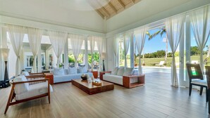 Ein geräumiges Wohnzimmer mit Holzboden, weißen Sofas und einem Couchtisch. Große Fenster mit transparenten Vorhängen bieten einen Blick ins Freie.