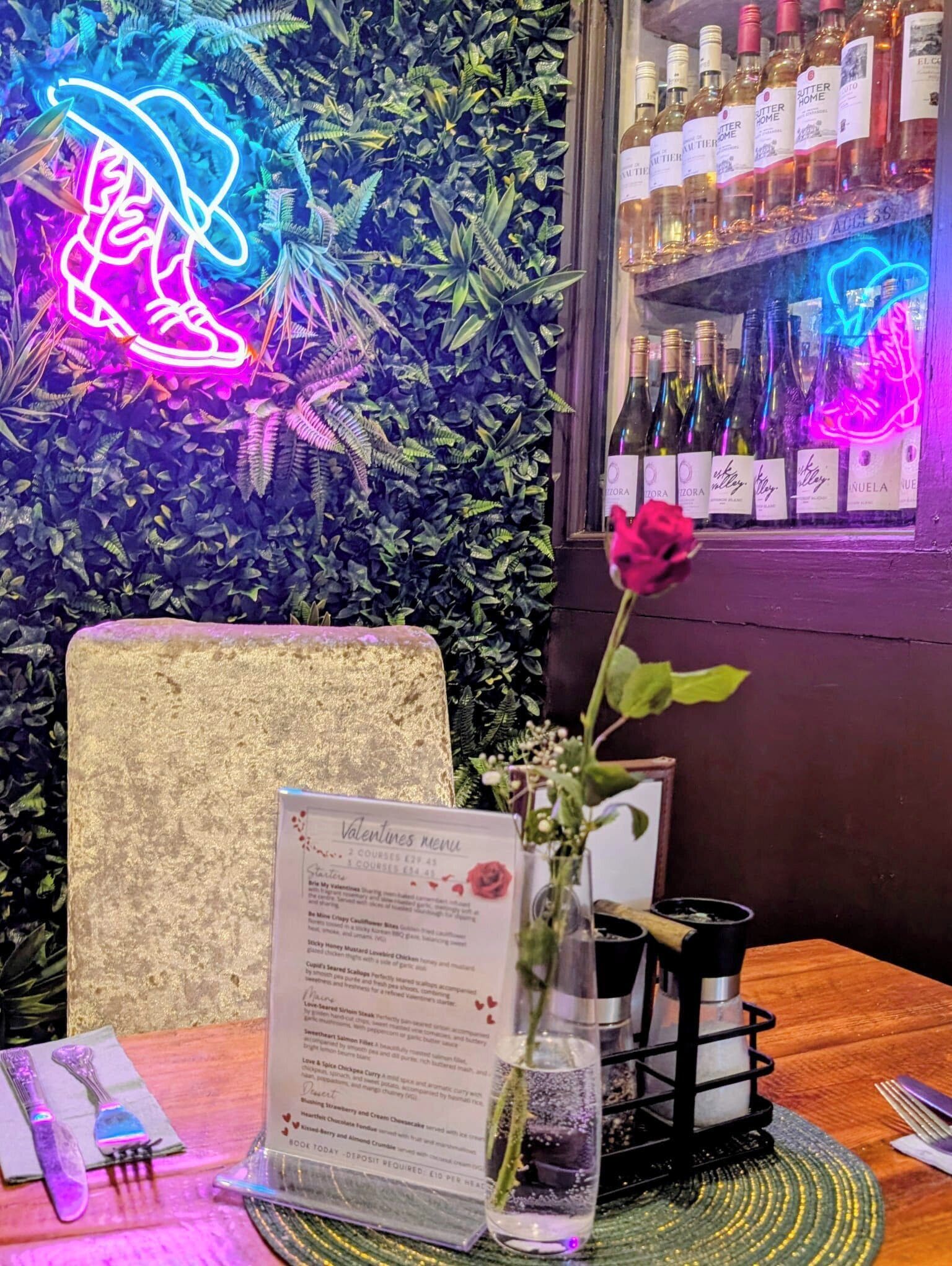 Et restaurantinteriør med et neonskilt, en menu, en rose i en vase og et vinskab.