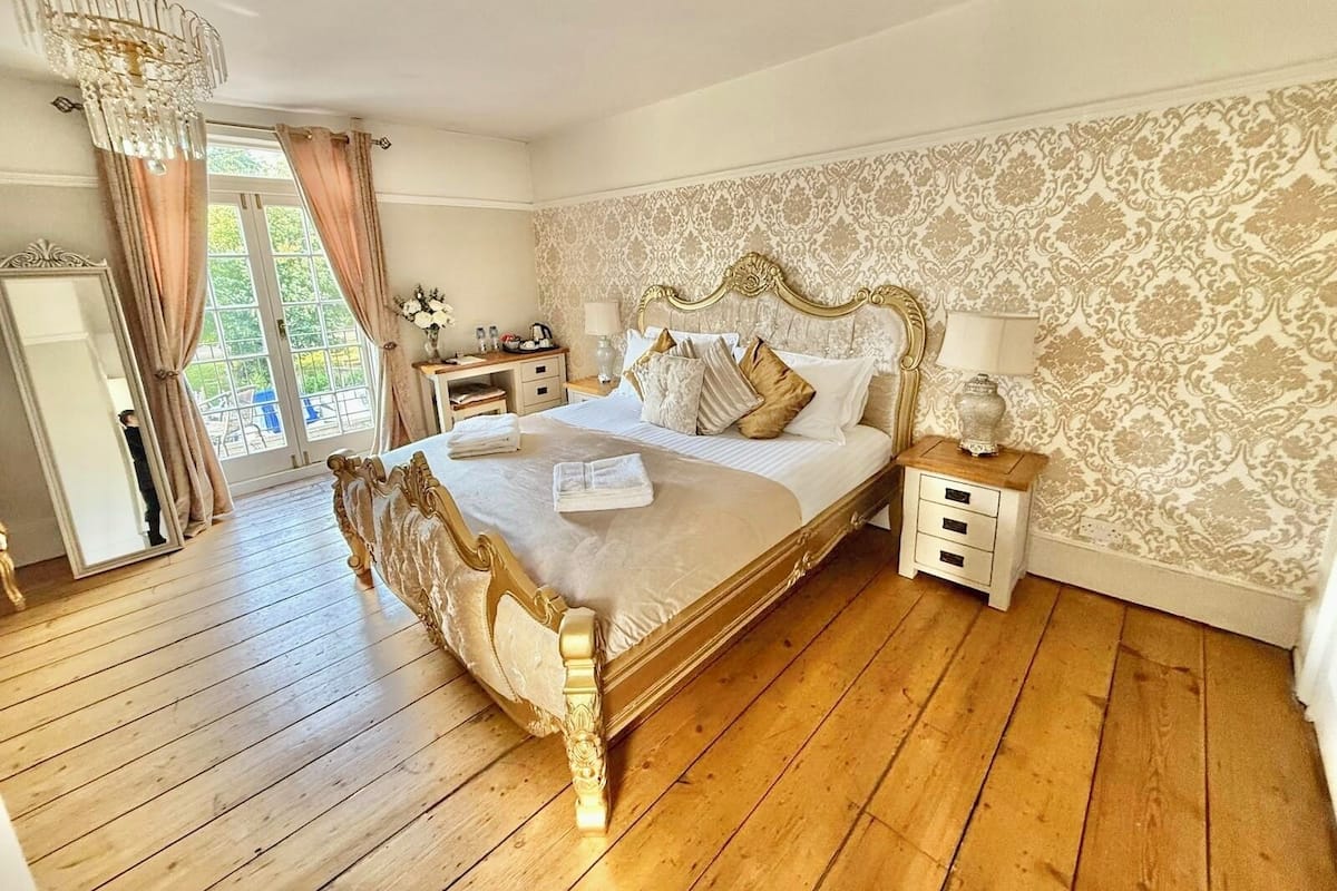 A spacious bedroom with a large bed, wooden flooring, a chandelier, and a view of the outdoors.