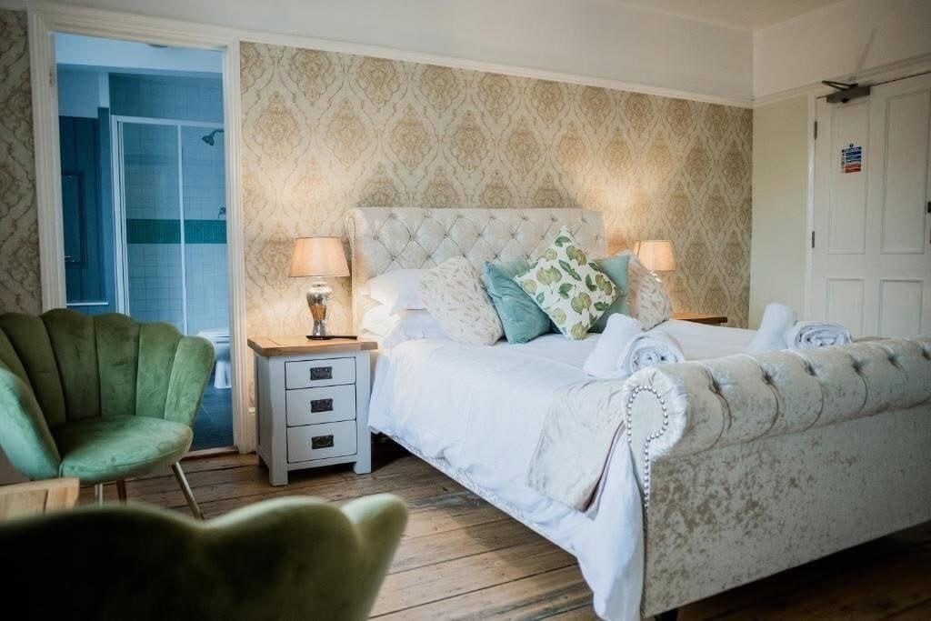 Une chambre à coucher avec un lit doté d’une tête de lit capitonnée, deux lampes de chevet, un fauteuil vert et un plancher en bois.