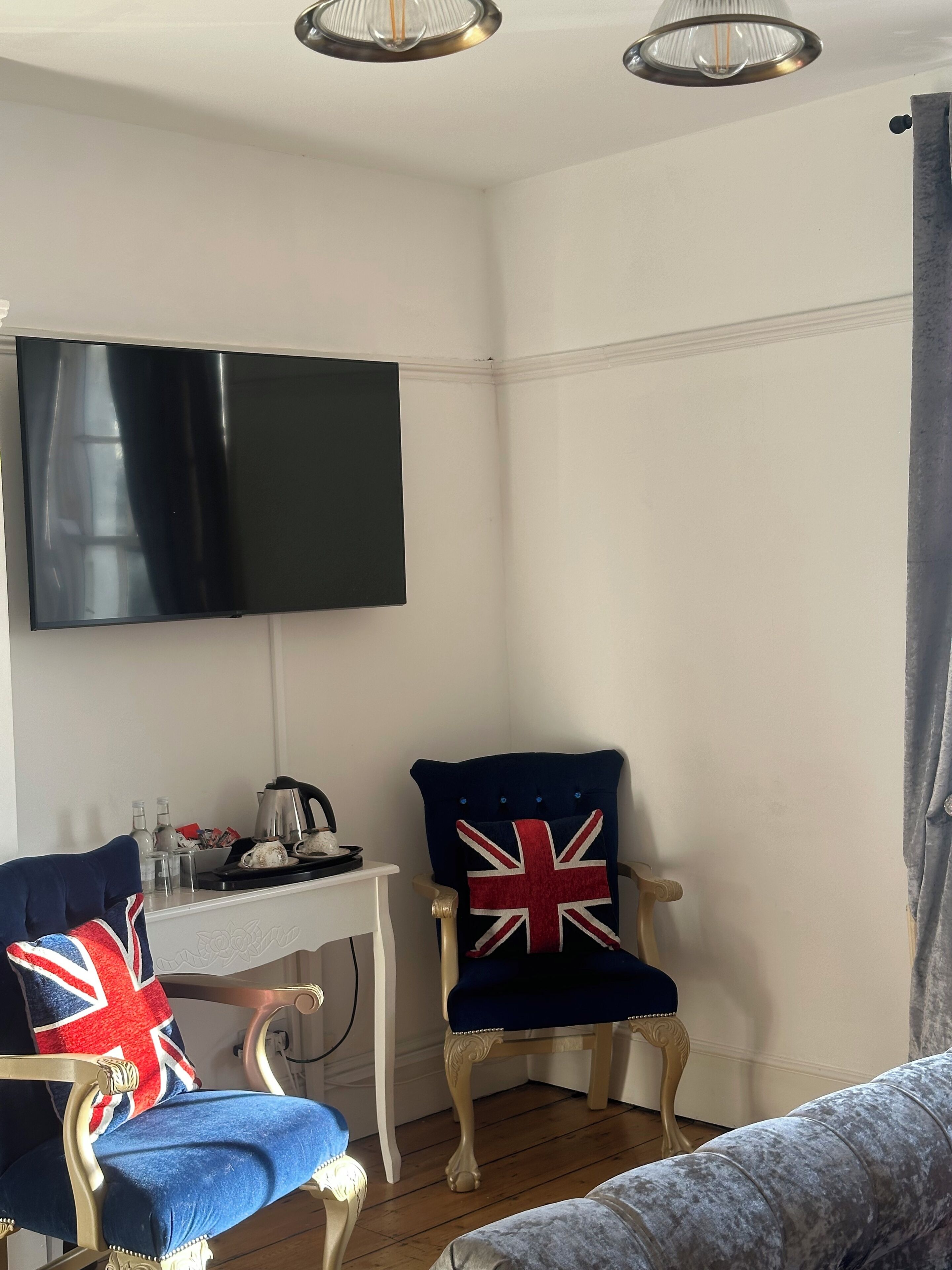 A room with a flat-screen TV mounted on the wall, a white table with a kettle and cups, two chairs with Union Jack pillows, and a grey sofa.