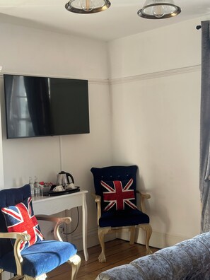 A room with a flat-screen TV mounted on the wall, a white table with a kettle and cups, two chairs with Union Jack pillows, and a grey sofa. - Grove Ferry Inn (Canterbury)