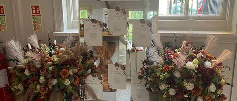 Eine Hochzeitslocation mit einem großen Spiegel, der den Raum reflektiert, floralen Dekorationen auf Ständern und einem Schild mit der Aufschrift 'The Owl's Wedding'.