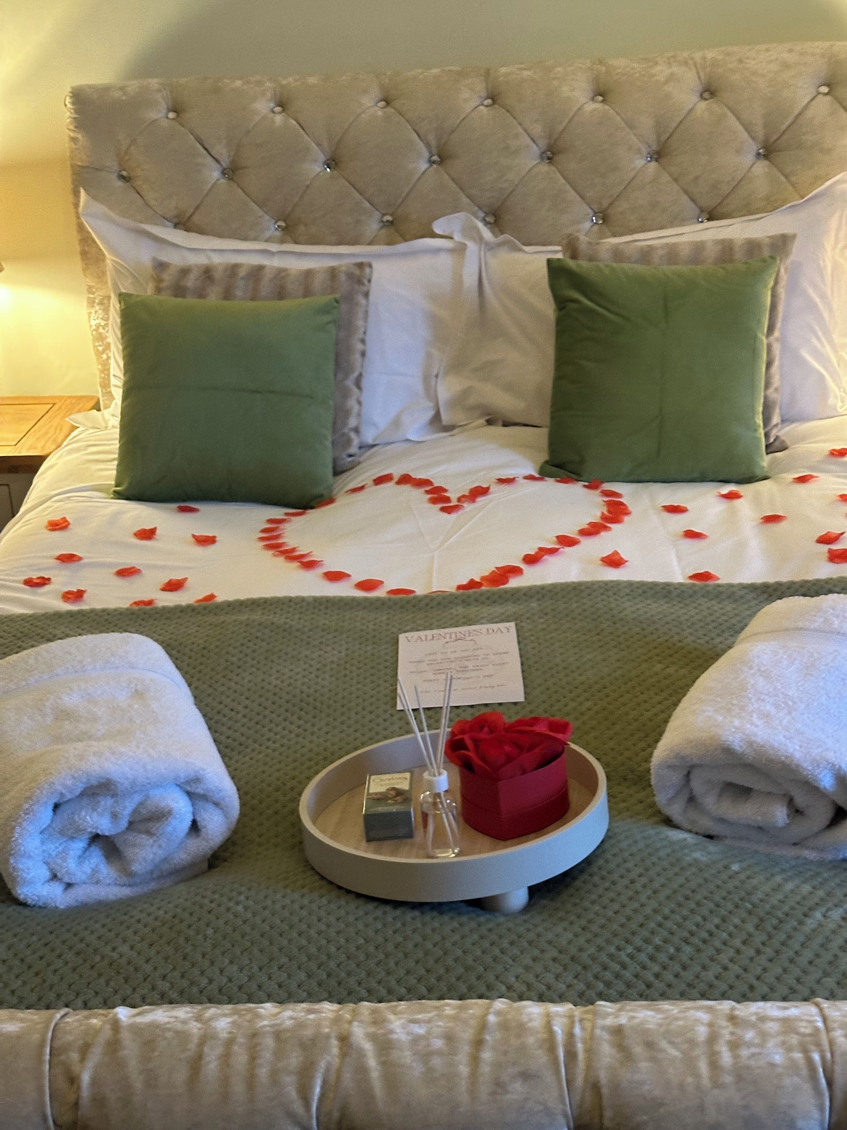 Quarto de hotel com cabeceira estofada, almofadas verdes e brancas, uma decoração em formato de coração na cama e duas toalhas ao lado.