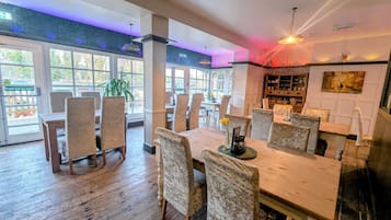 Ruime eetruimte met houten vloeren en beklede stoelen, grote ramen die natuurlijk licht binnenlaten, en een bar met planken en een schilderij.