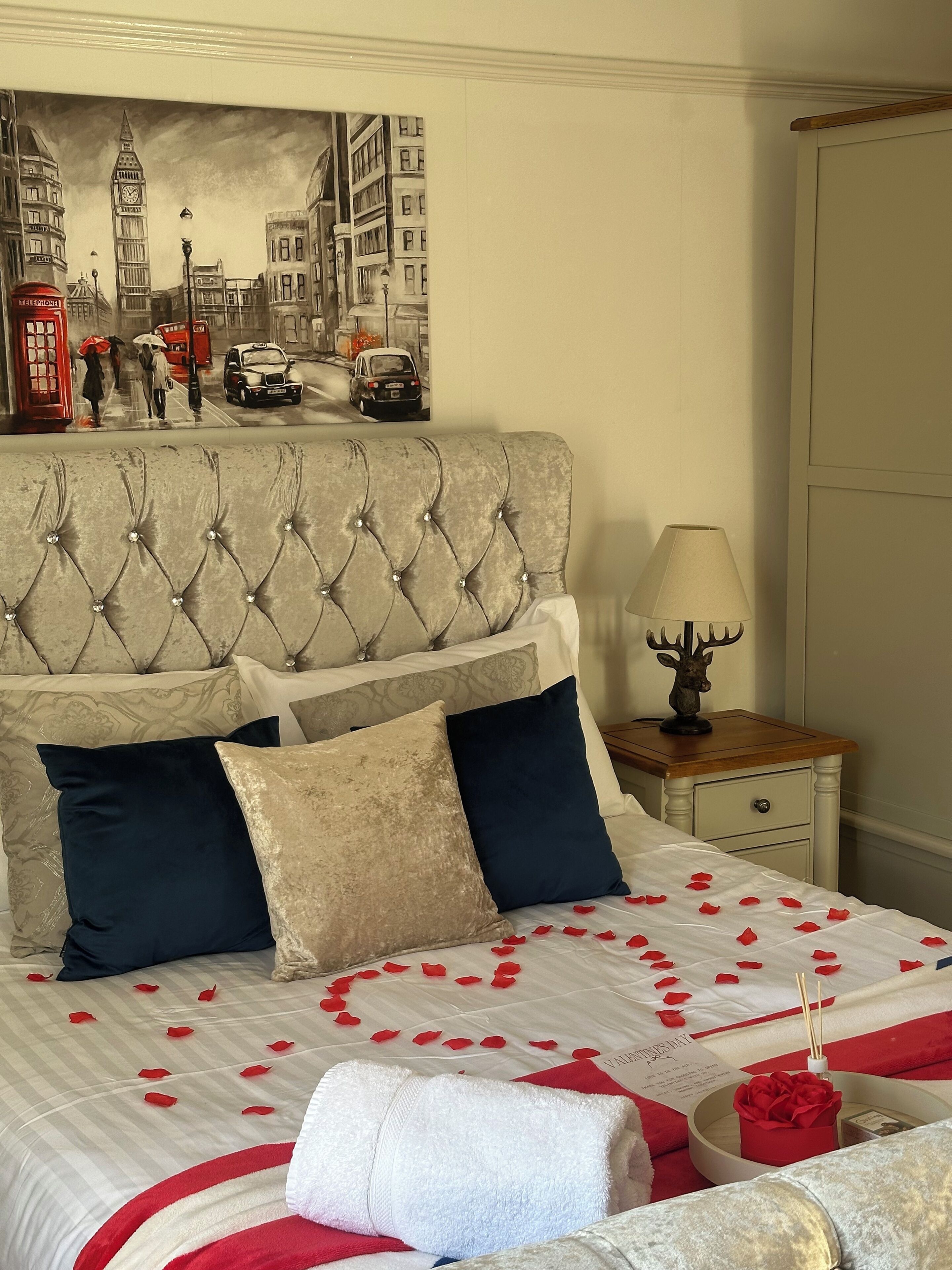 A bedroom with a tufted headboard, a bed with rose petals and a red rose, a lamp, and a painting of a cityscape.