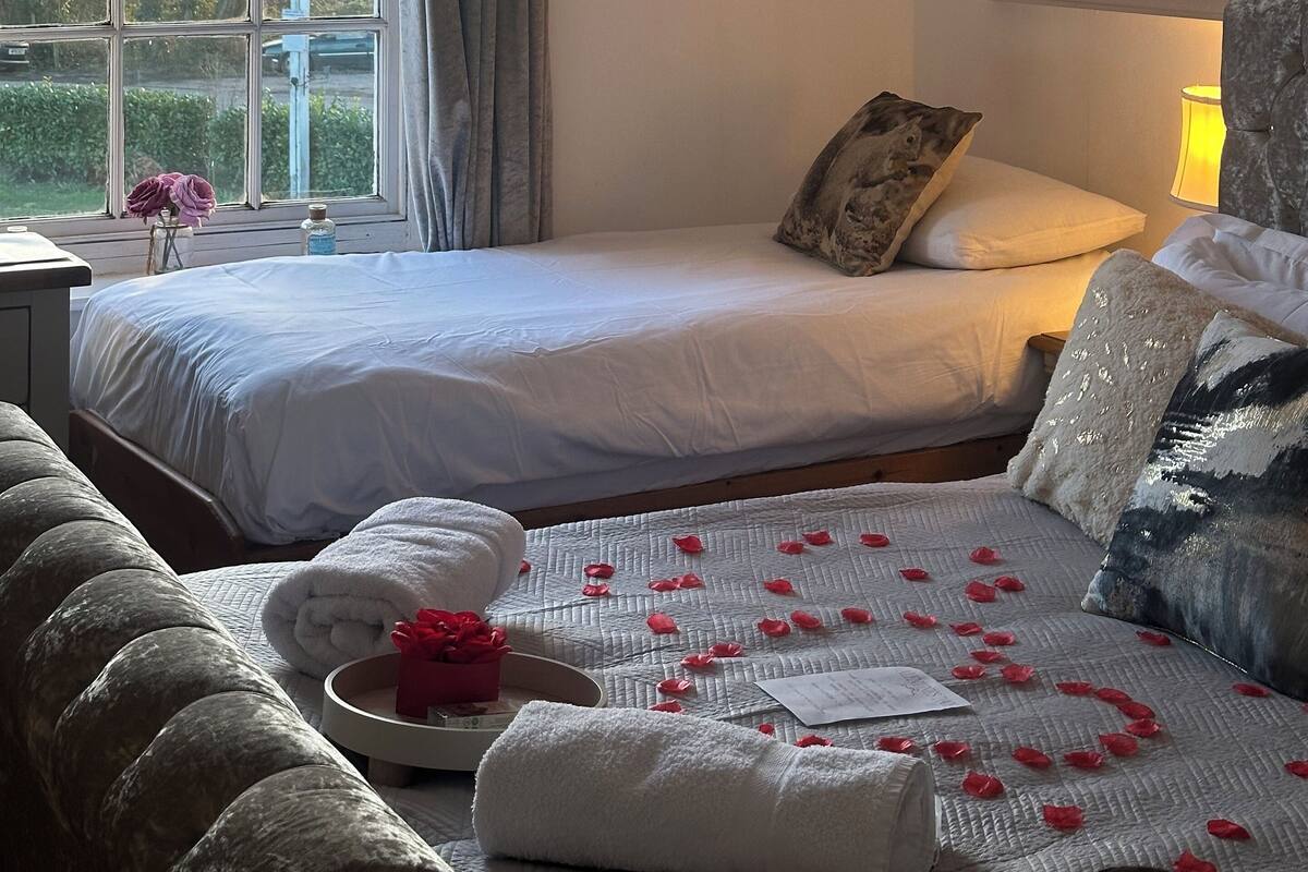 A hotel room with two beds, a large window with curtains, a painting on the wall, and a bedside table with a lamp.
