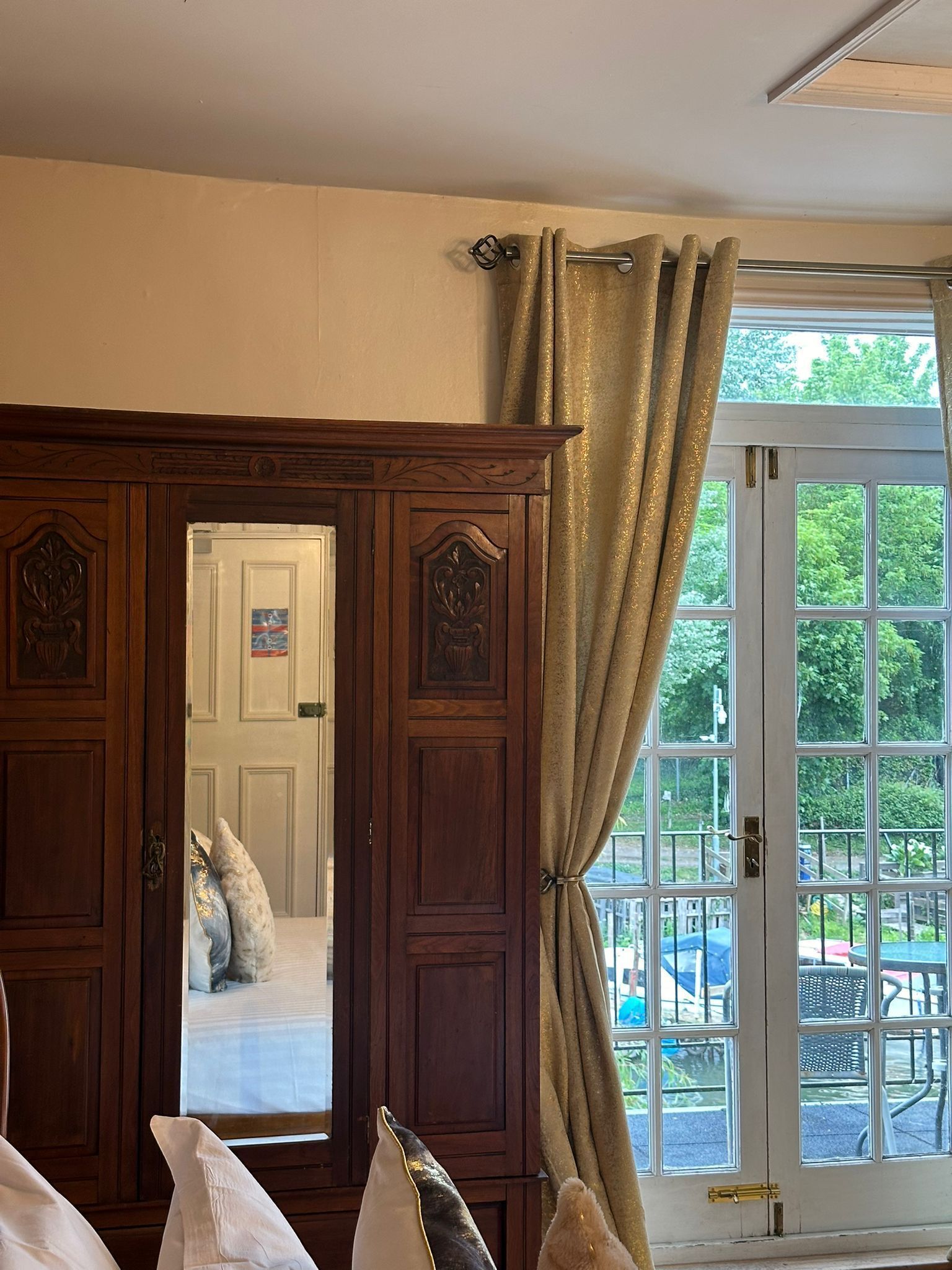 A room with a large wooden wardrobe, a bed with pillows, and a view of a garden through a glass door.