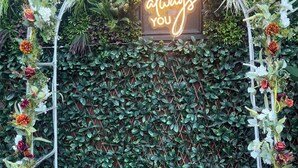 Een decoratieve boog met een neonbord waarop staat "It was always you", omgeven door weelderige begroeiing en bloemen.