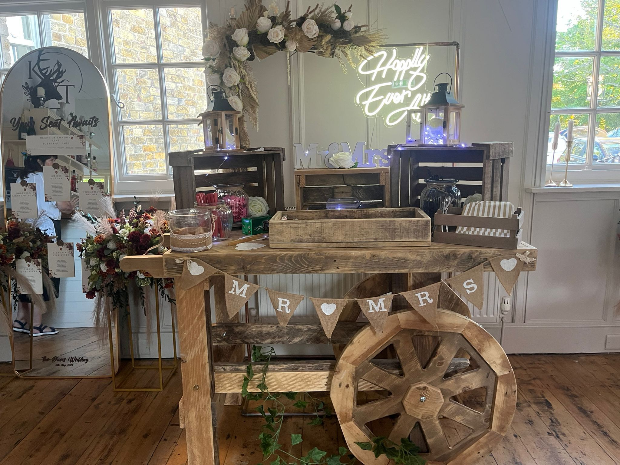 A rustic wedding setup with a wooden cart labeled "MR & MRS" and a neon sign saying "Happily Ever After".