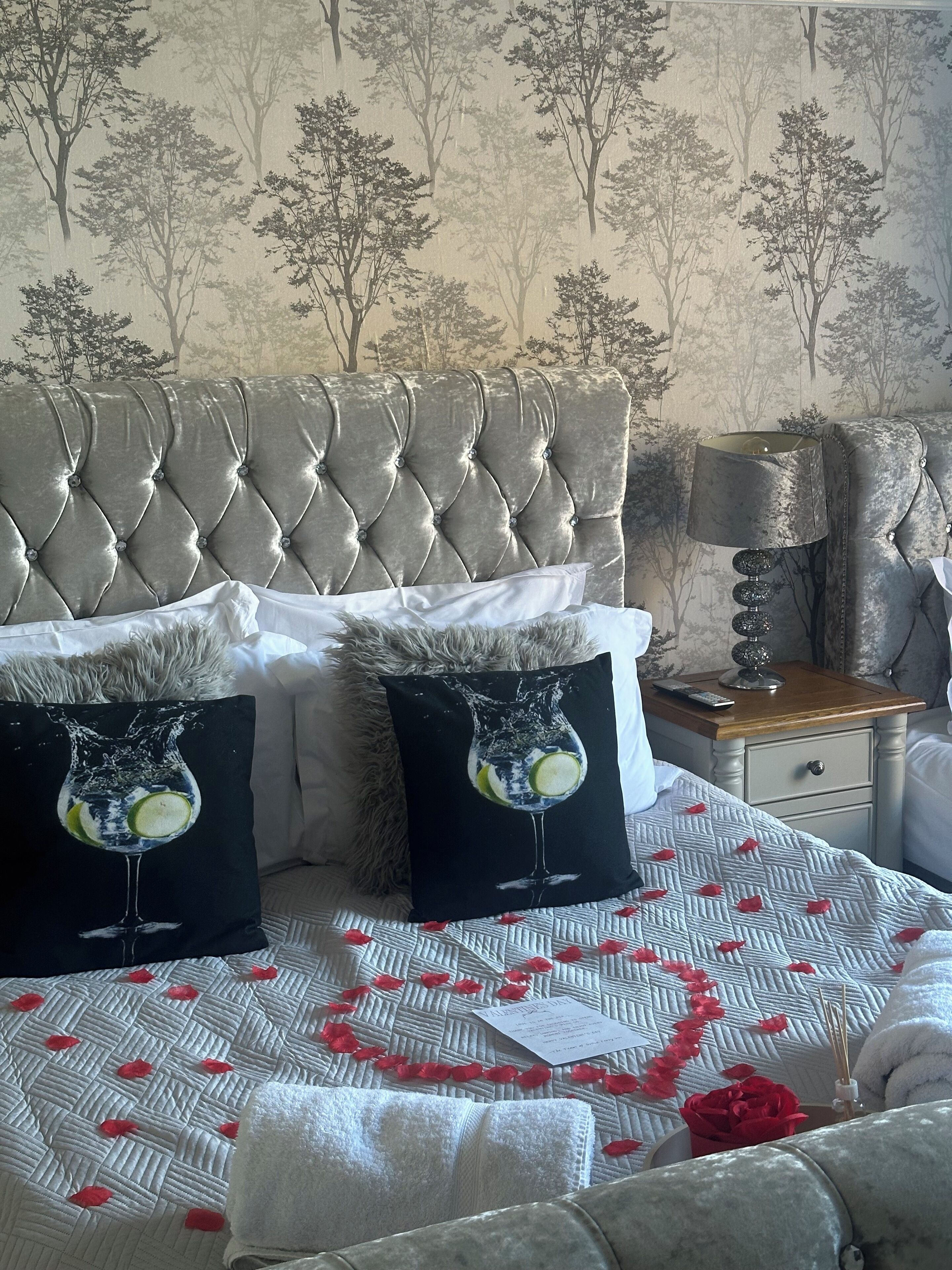 A bedroom with a tufted headboard, two decorative pillows, a bedside table with a lamp, and a bed with a quilted cover and rose petals.