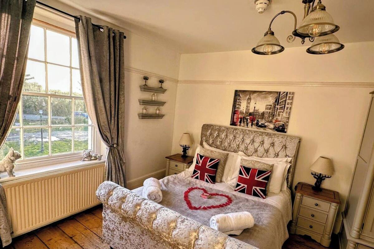 A bedroom with a large bed, a window with curtains, a radiator, a lamp, a nightstand, and a wall-mounted shelf.