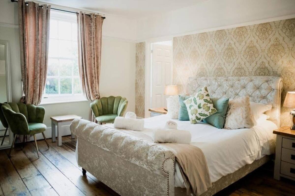 A bedroom with a large bed, two chairs, a nightstand, and a window with curtains.
