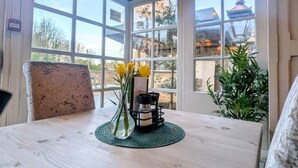 Een houten tafel met een vaas gele bloemen, een kandelaar en een groene placemat in een kamer met grote ramen en uitzicht op een gebouw met schoorsteen.